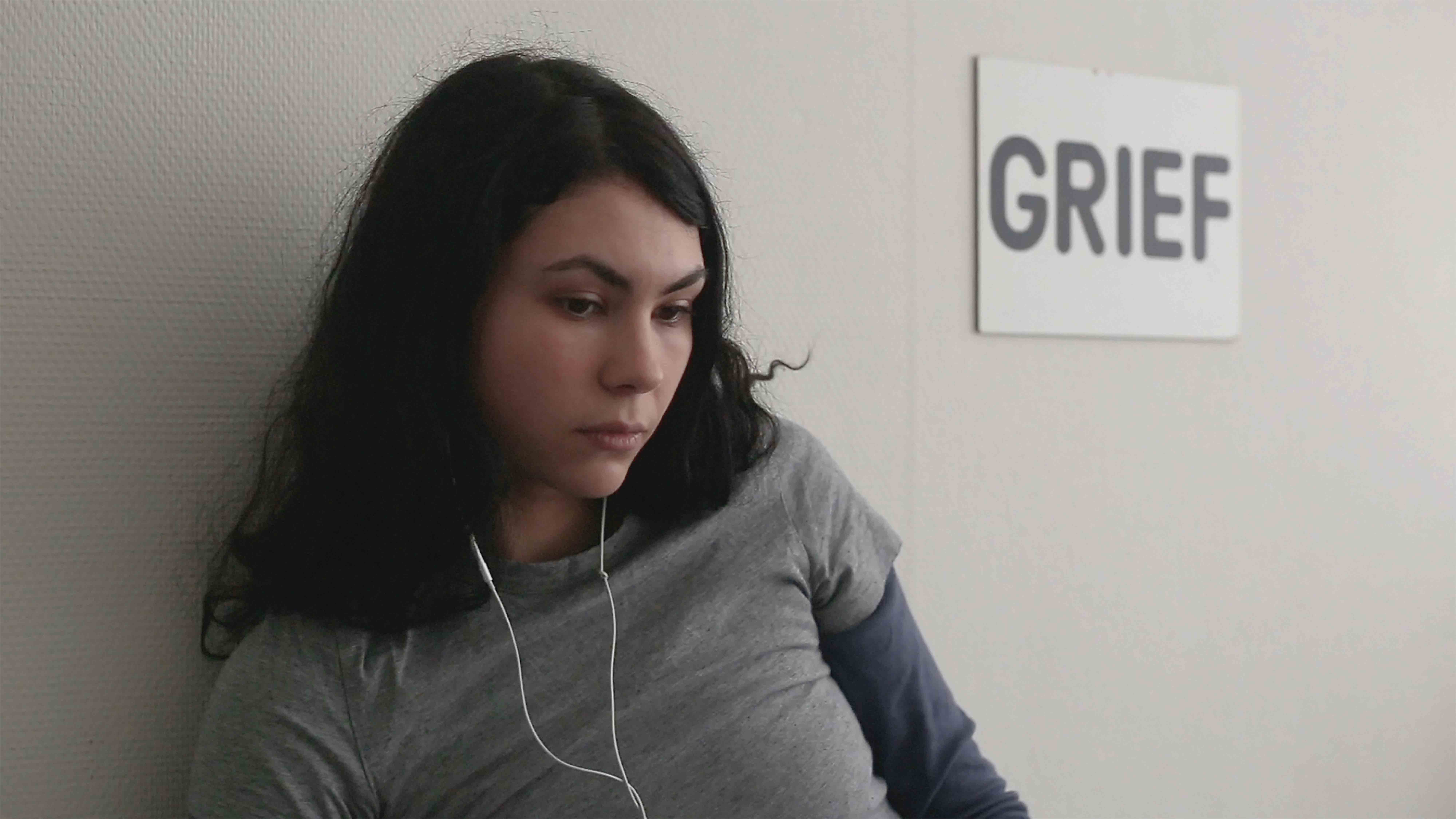 Woman with earphones in front of a sign reading "GRIEF", Grant Me Virtue But Not Yet