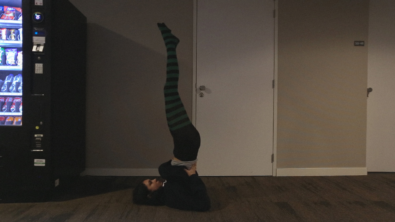 Person doing a shoulder stand in a dim hallway beside a vending machine, Grant Me Virtue But Not Yet